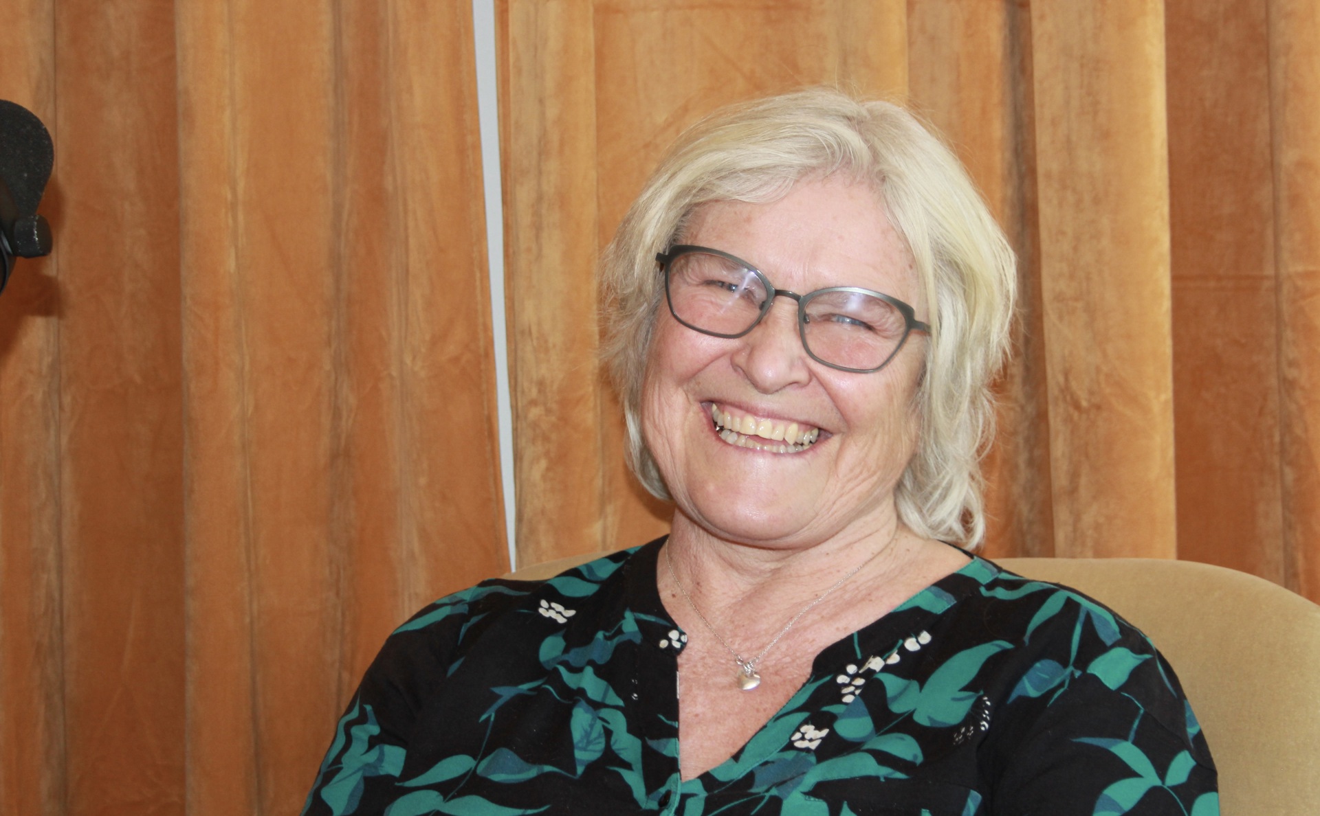 Smilende kvinne med lyst hår, briller og grønnmønstrete topp. Oransje fløyelsgardinger bak.