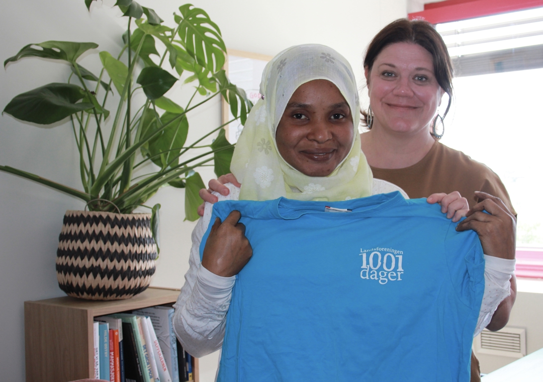 En melaninrik kvinne med hodeplagg og en hvit kvinne med mørkt hår. De holder en blå t-skjorte.