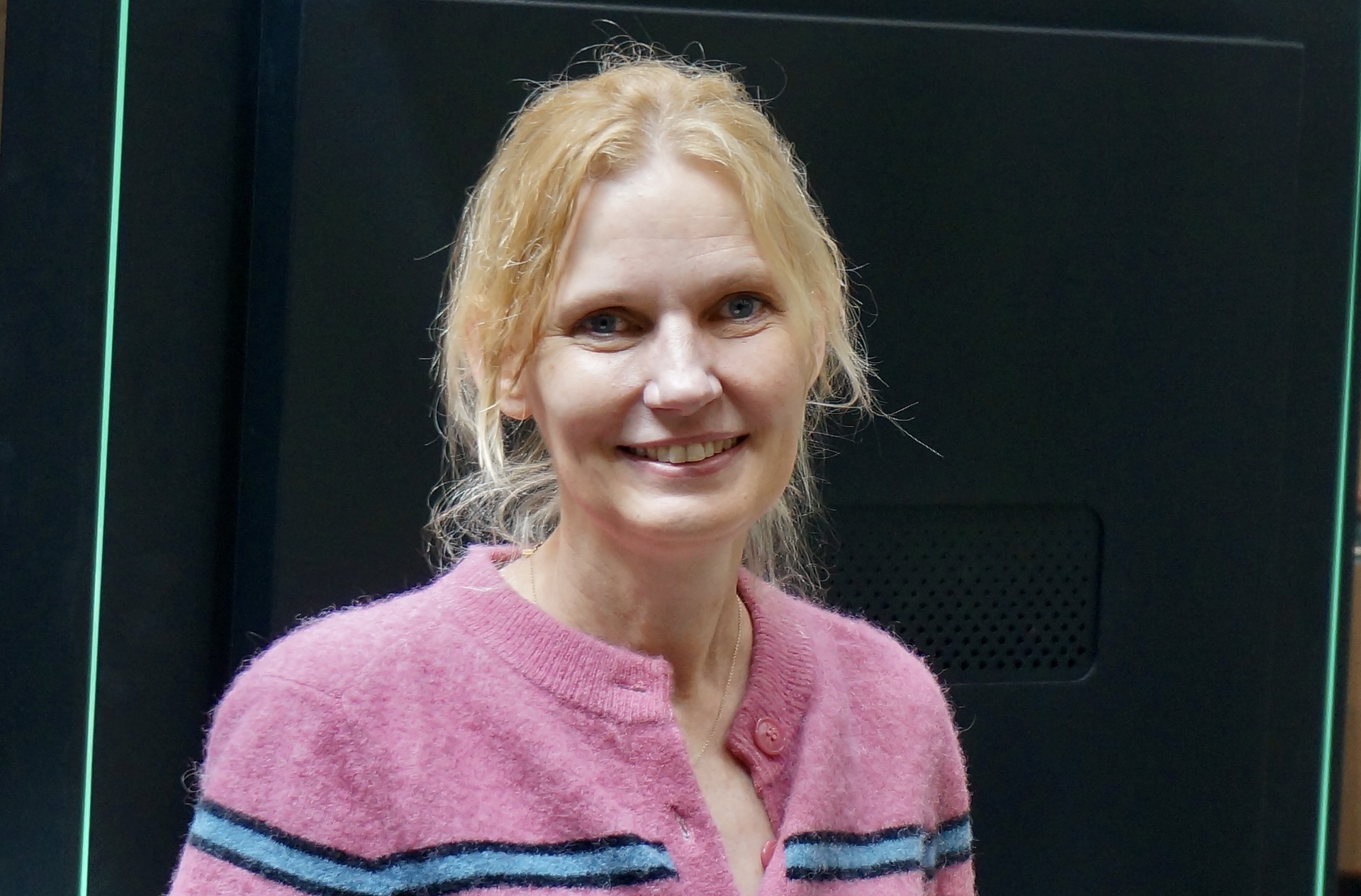 Kvinne, lyst hår, rosa strikkejakke med blå og svart stripe. Mørk bakgrunn.