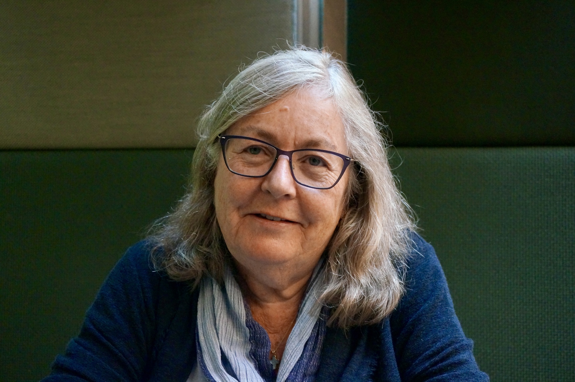 Kvinne med halvlangt grått hår og briller. Blå genser og lyseblått skjerf i halsen. Grønn bakgrunn.