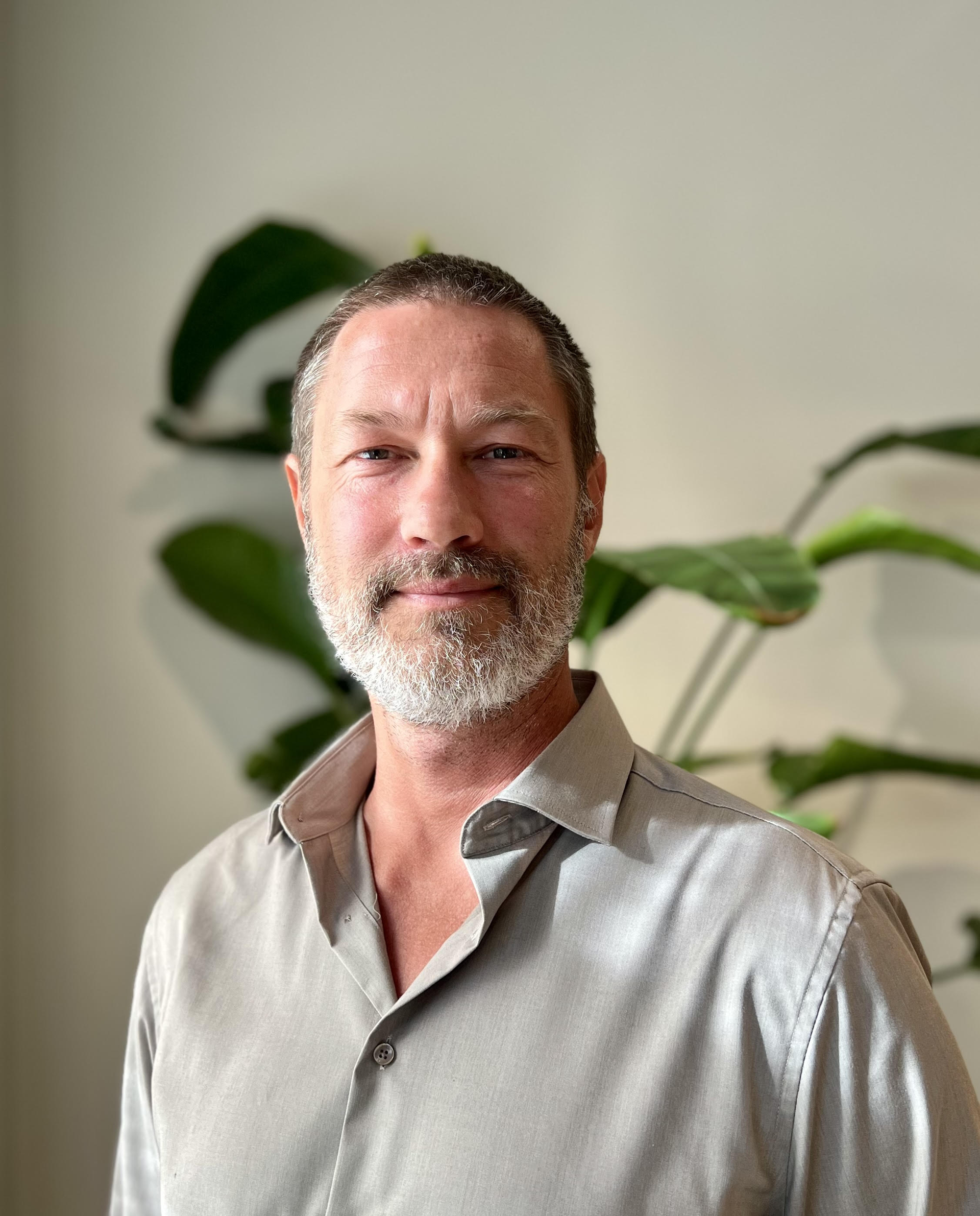 Mann med kortklipt hår, grått skjegg, grå skjorte. Bakgrunn lus med grønn plante