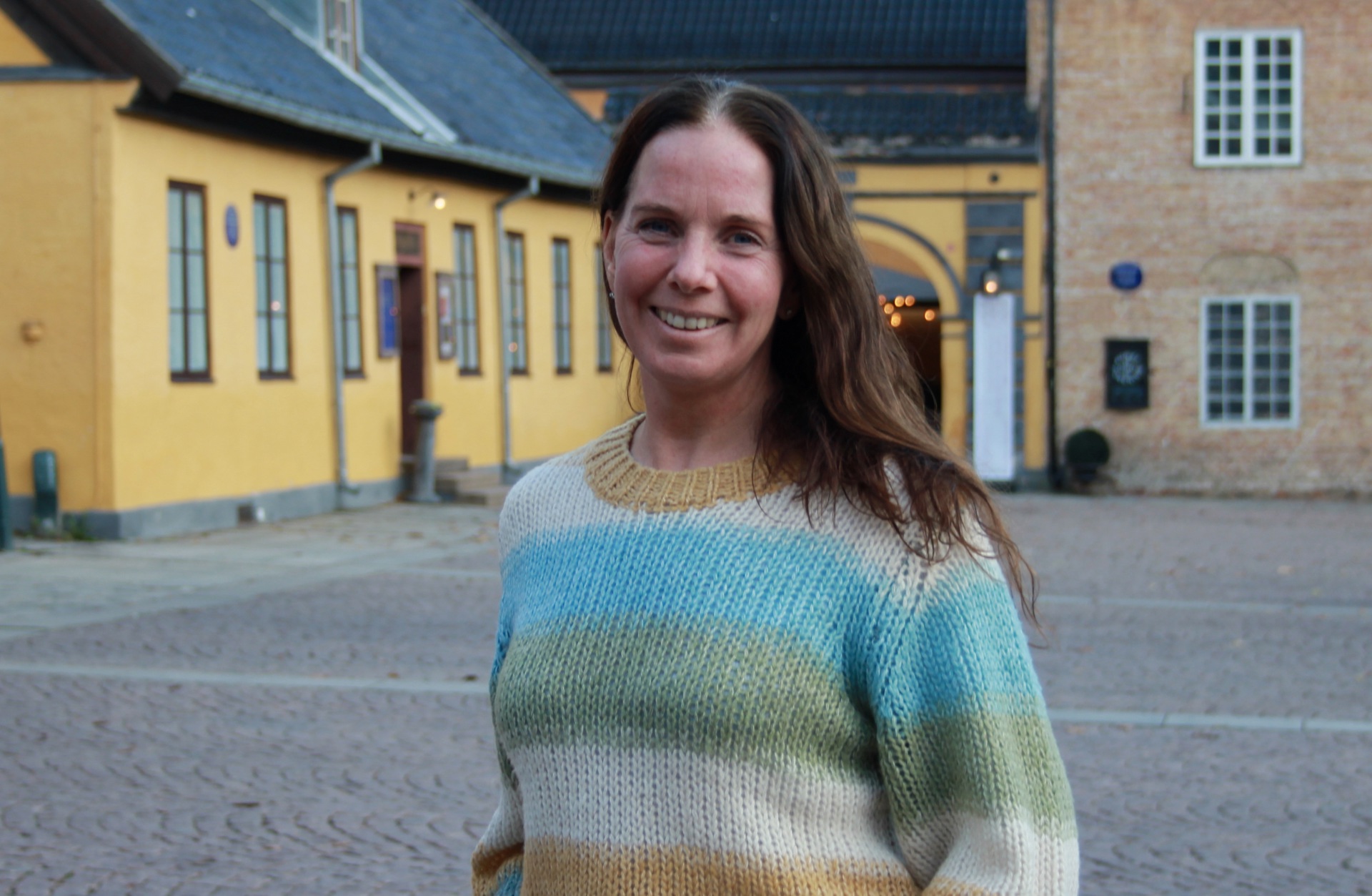 Smilende kvinne med langt brunt hår i fargerik genser utendørs på et torg. Gul bygning i bakgrunnen.