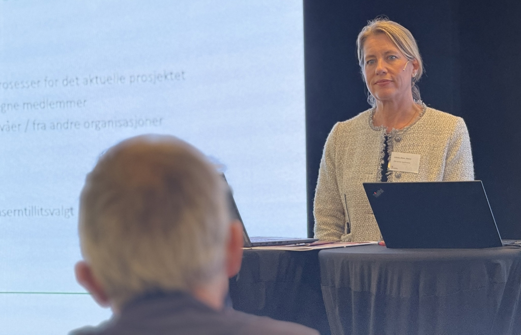 Kurset ble ledet av blant annet advokat Hanne Gillebo-Blom. Her står hun på talerstolen. Foran henne sitter en mann med grått hår, vi ser bare bakhodet hans og det er utydelig. Gillebo-Blomhar har lyst hår med sideskill og som er samlet i nakken. Hun ser konsentrert ut, og har på seg en lys jakke med sort topp inni.