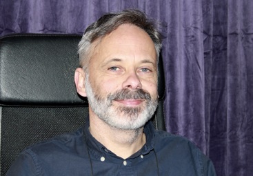 Mann, grått skjegg, mørkt/grått hår. Blå skjorte. Lilla fløyelsgardiner i bakgrunnen.