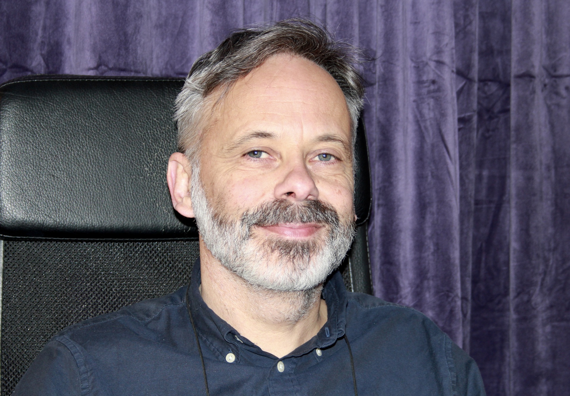 Mann, grått skjegg, mørkt/grått hår. Blå skjorte. Lilla fløyelsgardiner i bakgrunnen.