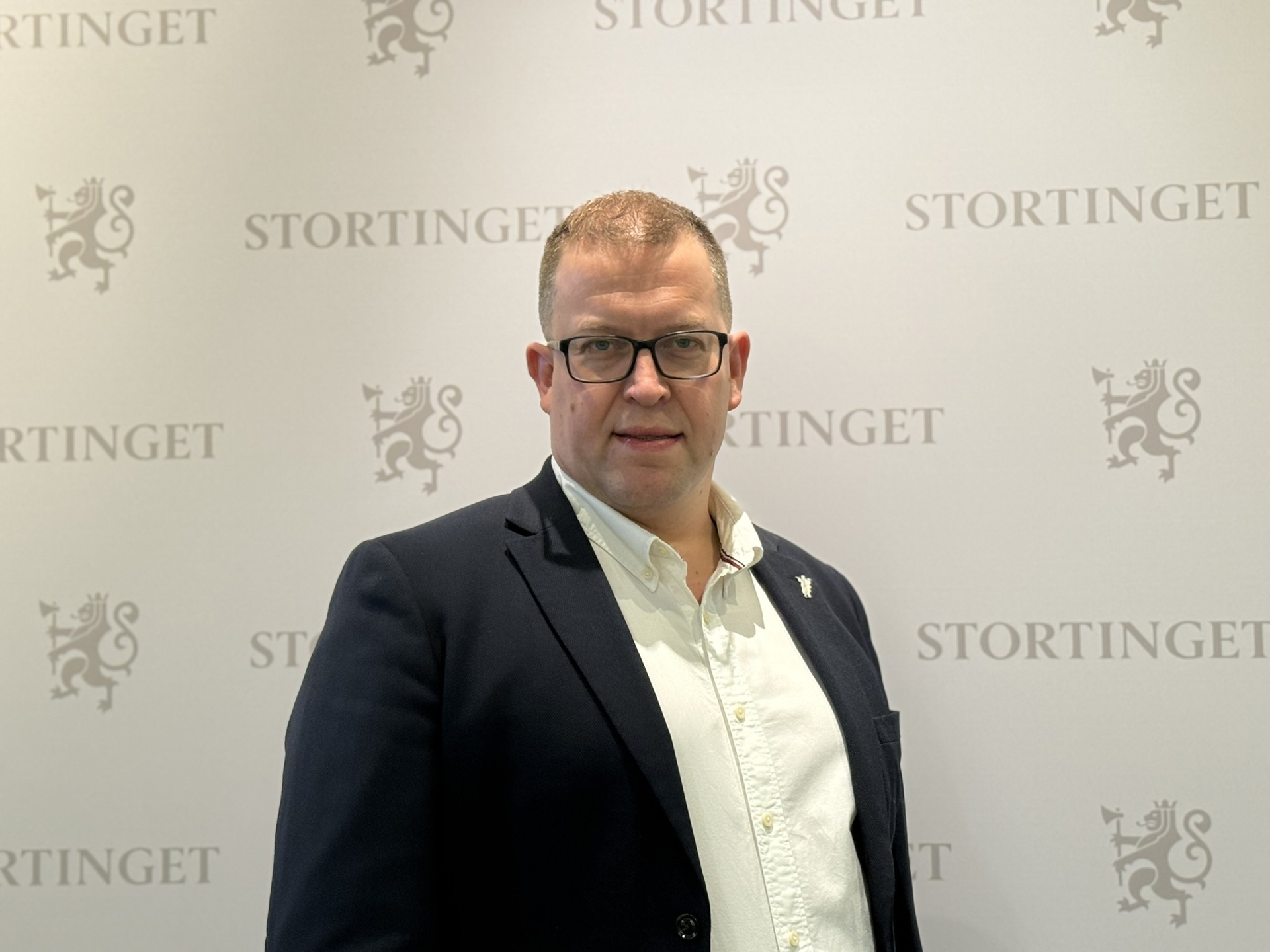 Leder for Allmenlegeforeningen, Hans-Christian Myklestul foran stortingets-logovegg. Han har kortklippet, lyst hår, firkantede briller med sort innfatning, har på seg sort dressjakke og hvit skjorte.