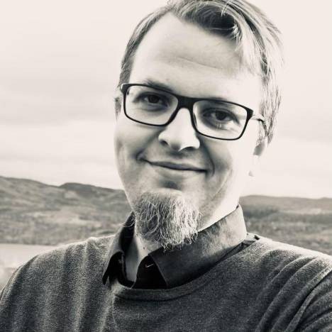 Sort-hvitt portrettfoto av en smilende mann med briller og skjegg.