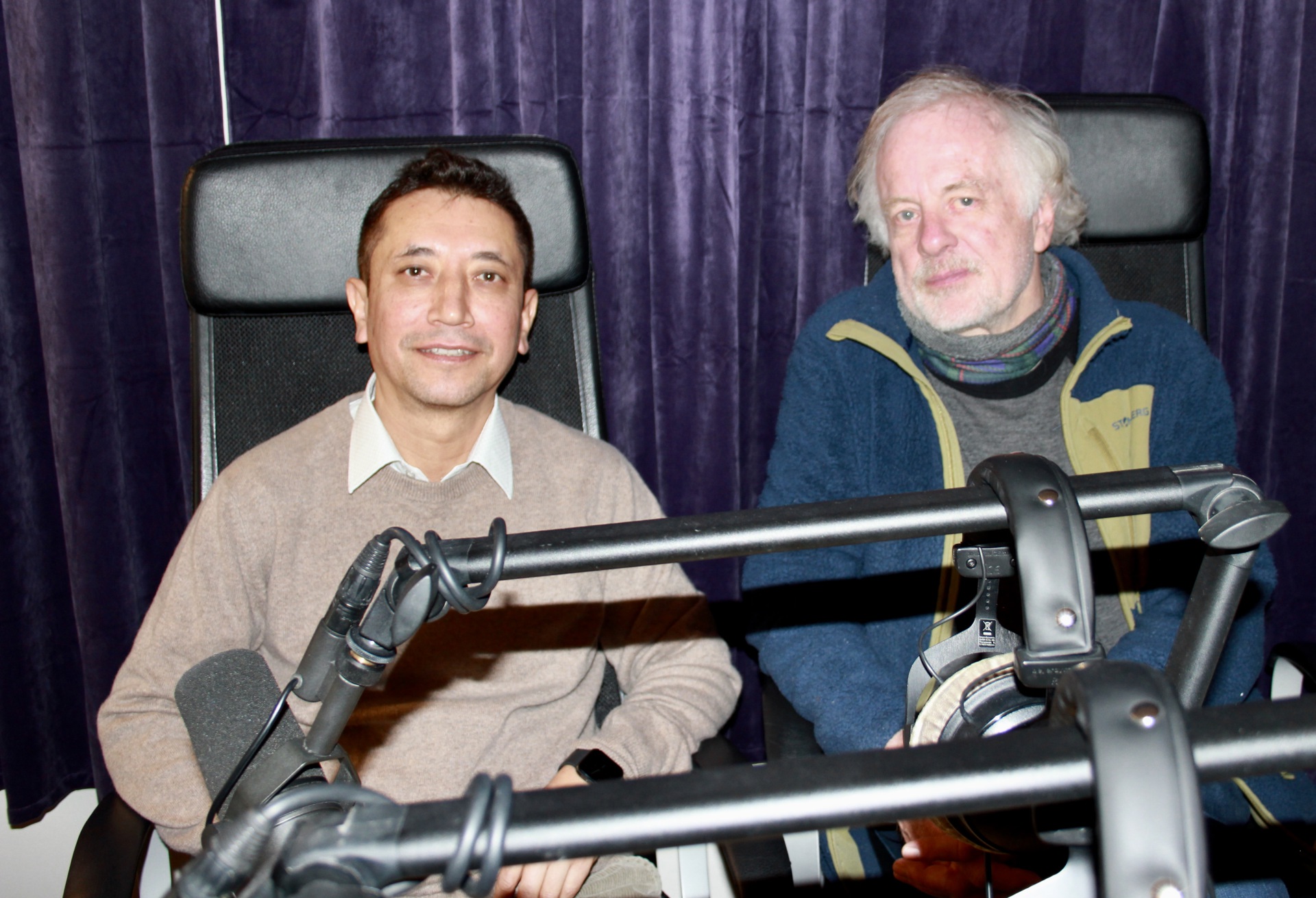 To menn bak mikrofonstativ. Mann til venstre i beige genser, hvit skjorte, mørkt hår. Mann til høyre i blå jakke med grønne detaljer, grå genser, grått hår.