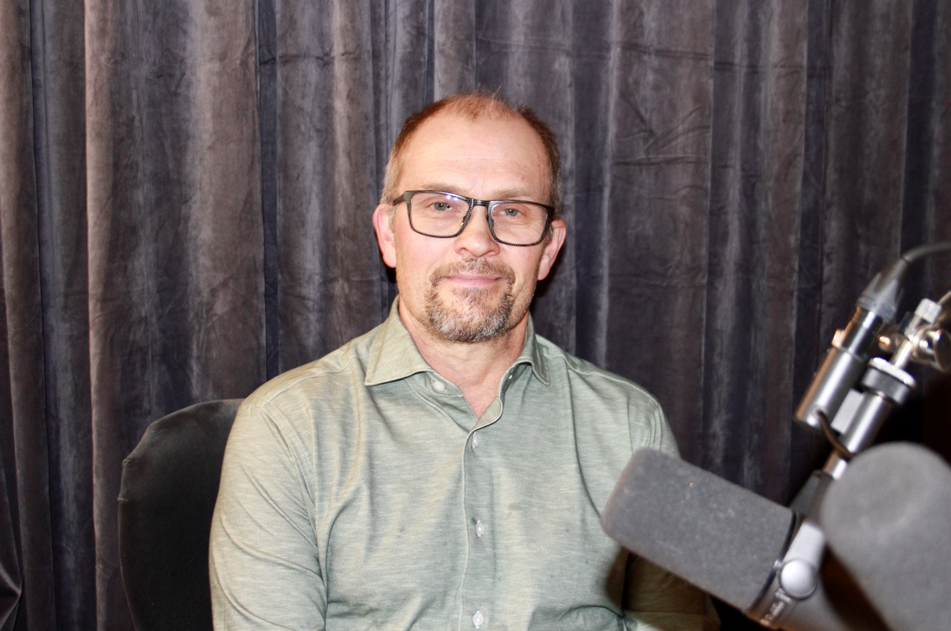 Mann med grå skjorte, skjegg, briller. Mikrofonstativ til høyre. Lilla fløyelsgardiner i bakgrunnen.