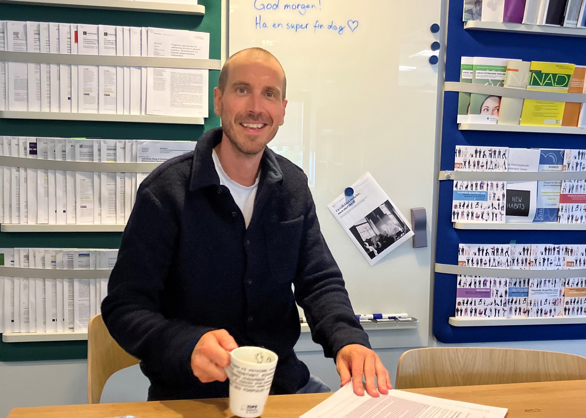 Blid mann sitter ved bord på kontor og holder en kaffekopp og et ark