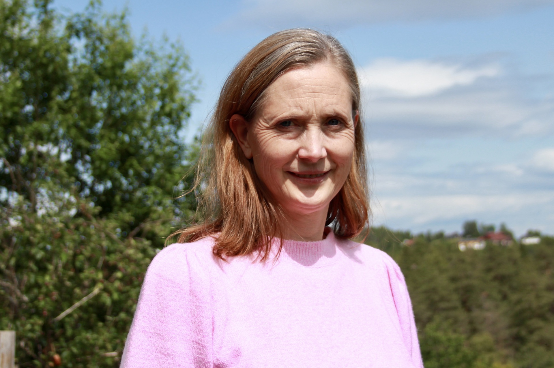 Kvinne med langt, blondt hår. Rosa genser. Grønt bladverk og blå himmel i bakgrunnen.