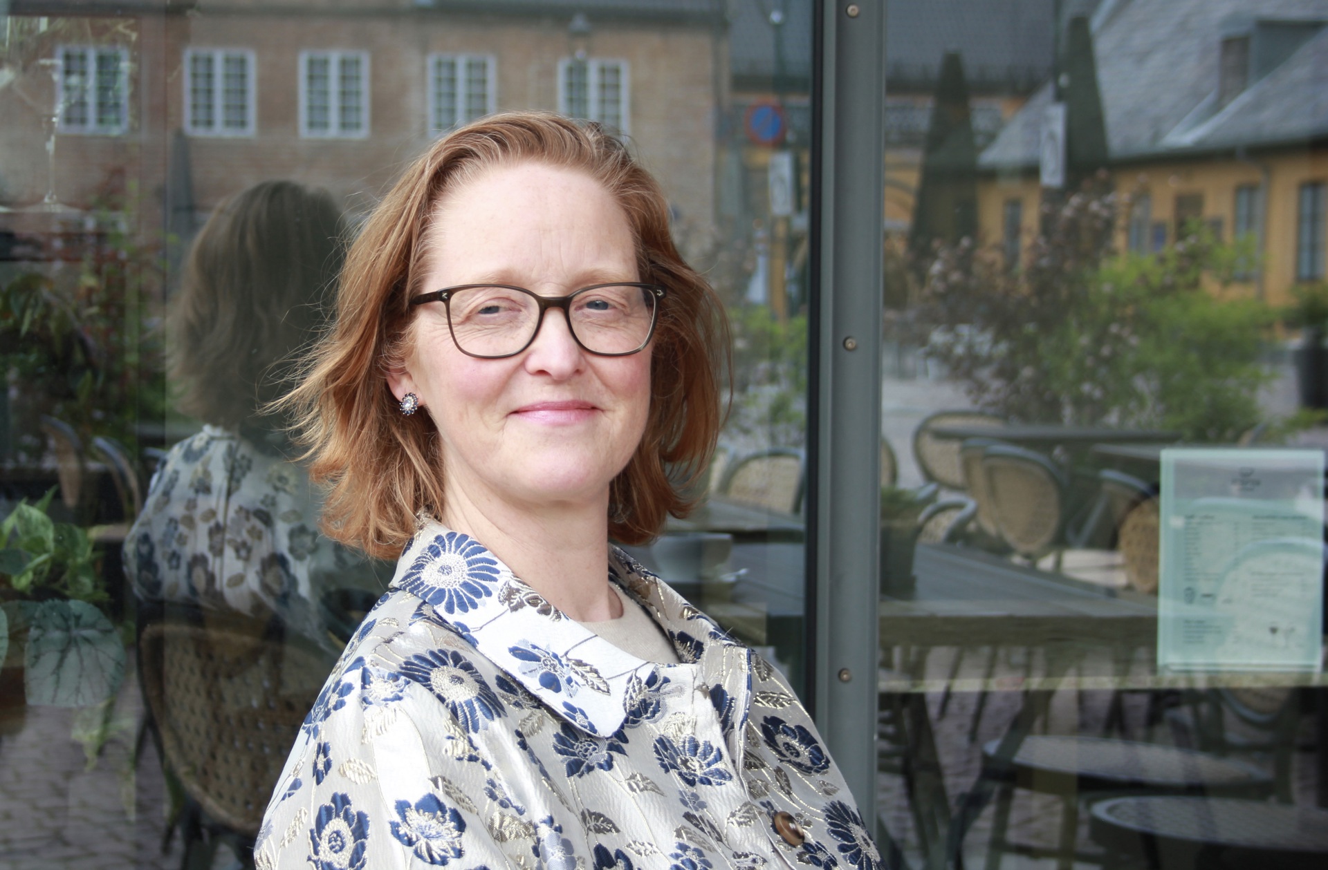 Kvinne med halvlangt, blondt hår, briller. Lys topp med blå blomster. Vindu i bakgrunnen.