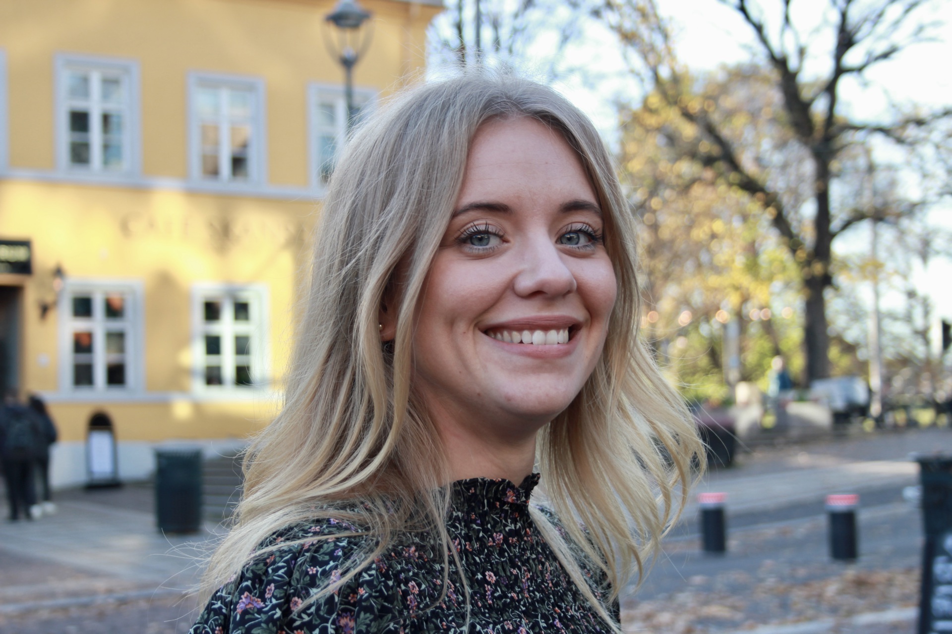 Ung smilende kvinne med langt lyst hår. Utendørs. Gul murbygning i bakgrunnen.