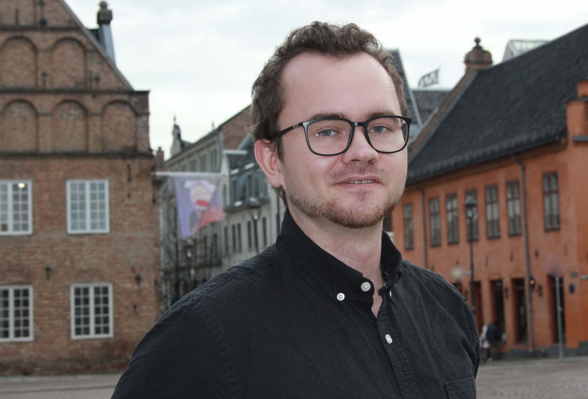 En ung mann med briller, skjegg og svart skjorte utendørs på en plass omkranset av murbygninger.
