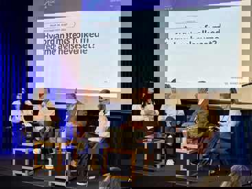 Fire personer i panel diskusjon på scene foran skjerm