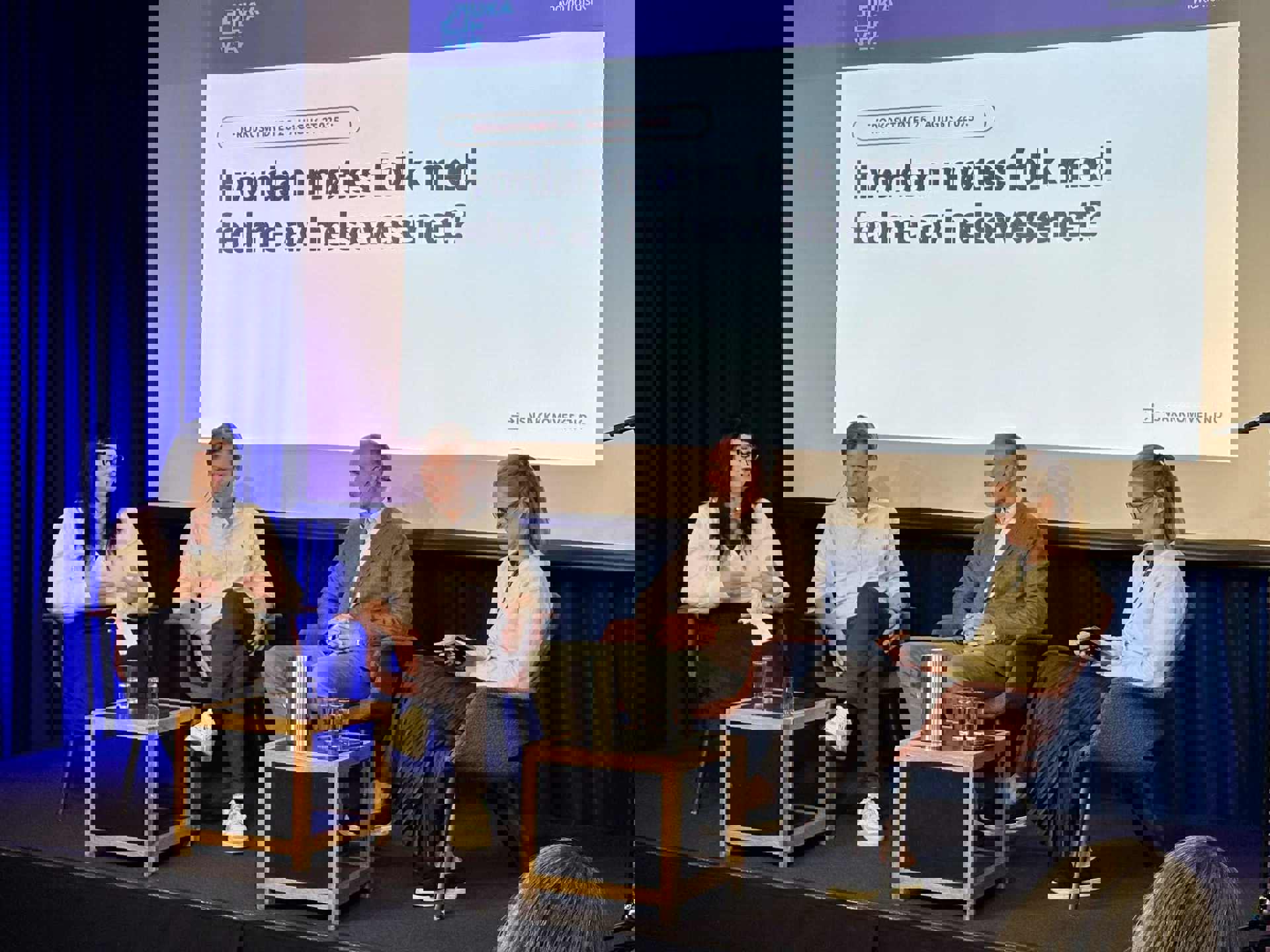 Fire personer i panel diskusjon på scene foran skjerm