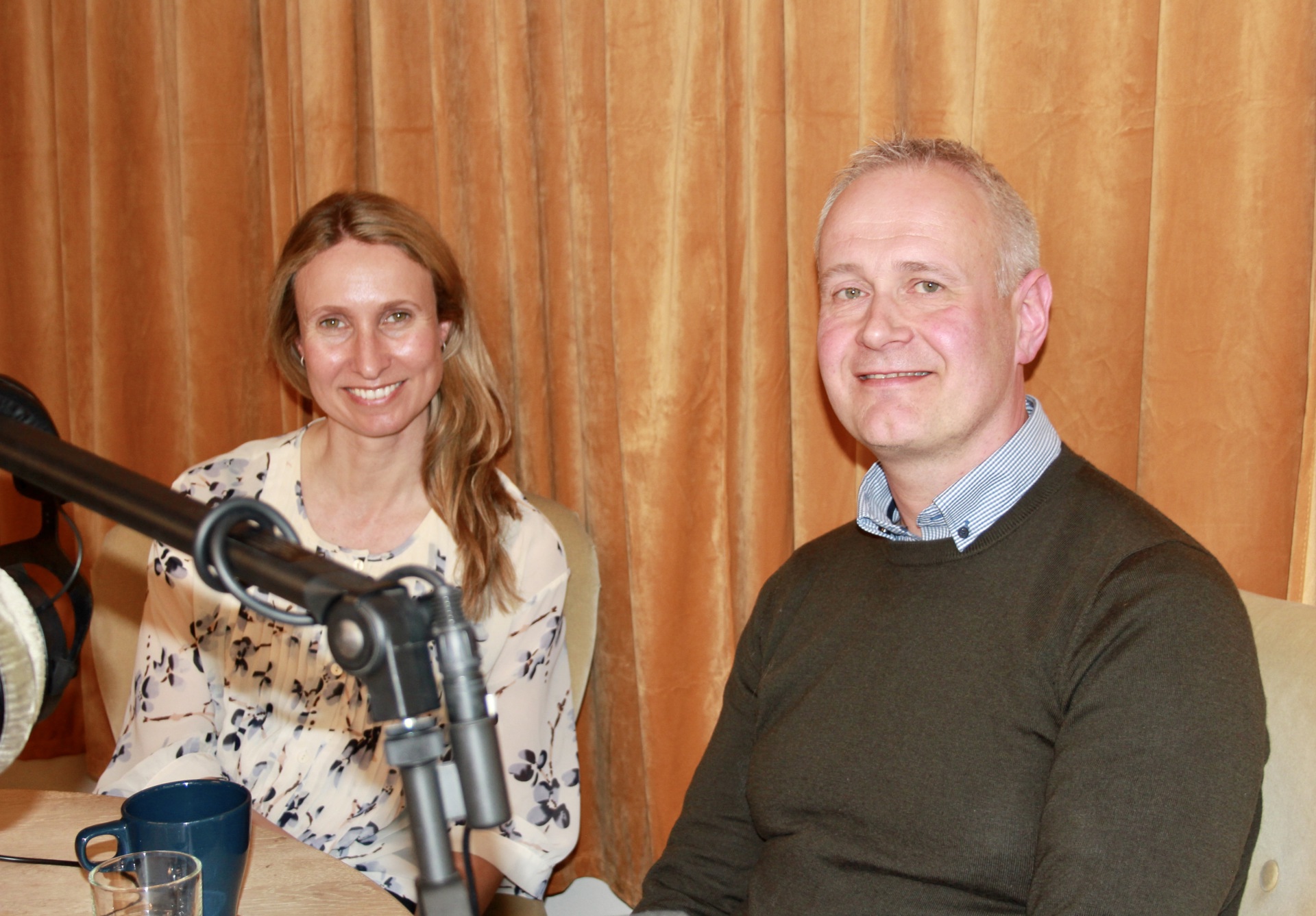 Kvinne med lyst, langt hår og lys, blomstrete topp. Mann med lyseblå skjorte i mørk genser. Mikrofoner. Gule fløyelsgardiner i bakgrunnen.