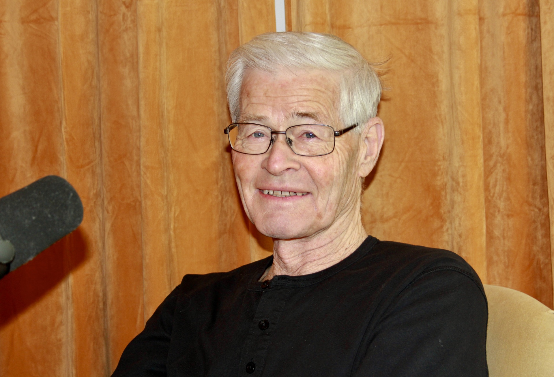 Mann, briller, grått hår, smiler. mørk genser. Mikrofon til venstre. Gule gardiner bak.