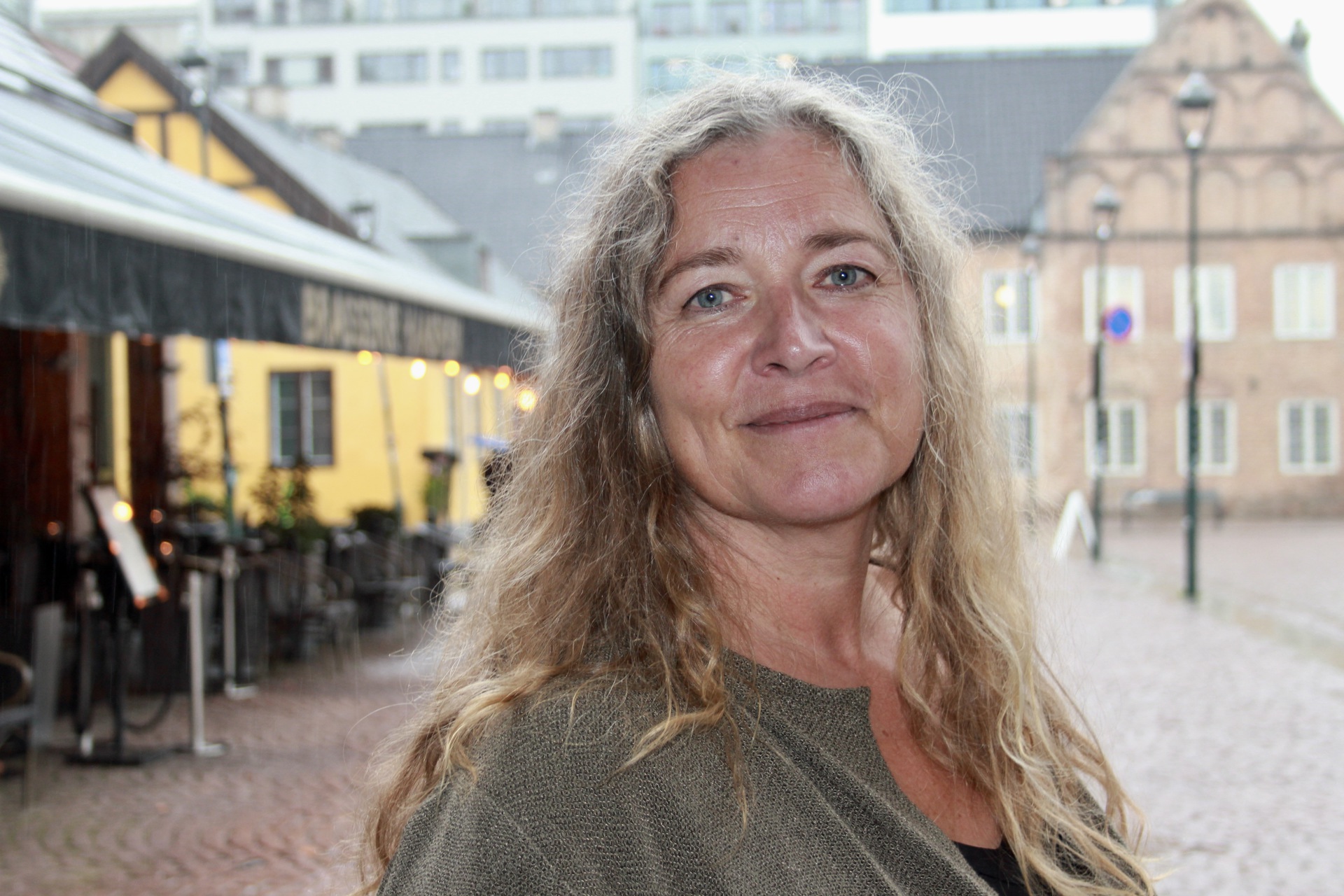 Bilde av kvinne med langt lyst hår på en åpen plass i byen.