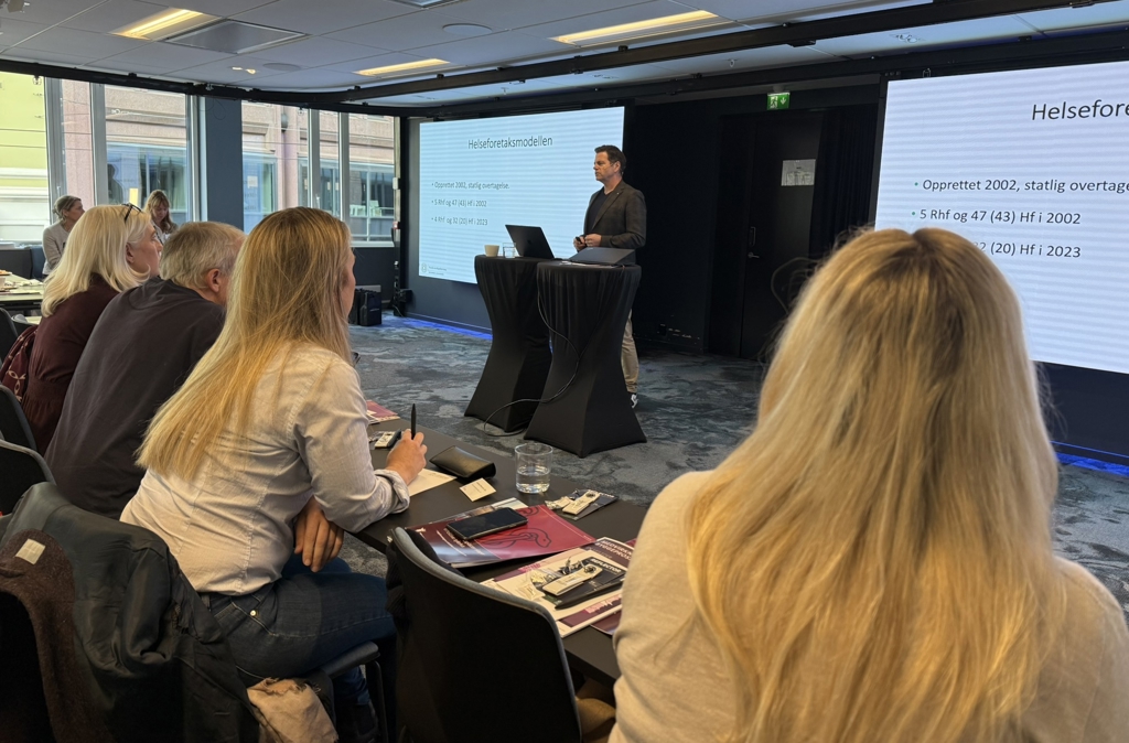 Ståle Clementsen, leder i Overlegeforeningen holder foredrag om Helseforetaksmodellen. Foran ham sitter en rekke mennesker som vi ser bakfra. Han står bak en sort talerstol, han har brunt, kortklippet hår og grå dressjakke med sort genser inni. Han har lyse bukser.