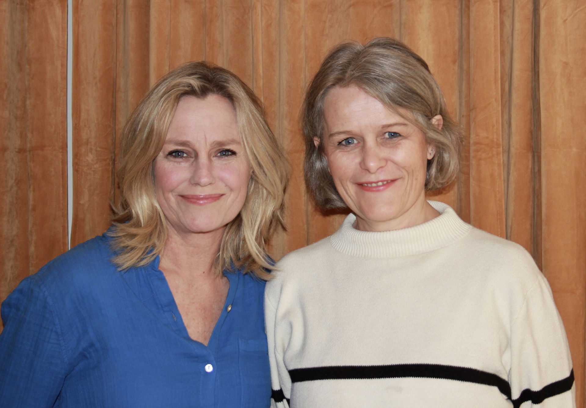 To kvinner foran en gul gardin. En med lyst hår og blå topp, en med blondt hår og hvit genser med svart stripe.